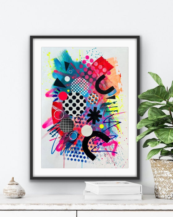 A black framed vibrant, colourful abstract artwork by artist Kate Elkington, leaning on a shelf with a book and coffee cup, featuring bold, graphic shapes, dynamic brushstrokes, and playful patterns. The composition includes splashes of neon pink, orange, bright red, and blue, layered with geometric elements such as dots, grids, and curved black lines, radiating energy and movement.