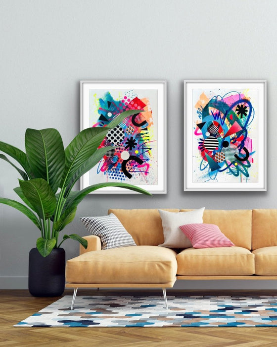 An interior with a yellow sofa, green plant and on the wall two large, white framed, vibrant abstract artworks by Kate Elkington featuring a dynamic mix of bold shapes, patterns, and colours. The composition includes overlapping circles, triangles, and lines in neon pinks, blues, oranges, and yellows, accented with stripes, polka dots, and grid designs. The pieces radiate energy and playful creativity.