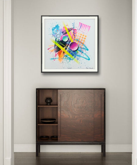 An interior with a dark wood cabinet. Hanging above it in a black frame is a vibrant abstract artwork by Kate Elkington bursting with neon hues of pink, orange, yellow, and blue. The composition features bold circular shapes, polka dot patterns, stripes, energetic paint splatters, and expressive brushstrokes. Layered textures add contrast and depth, creating a dynamic piece full of playful movement and joyful energy.