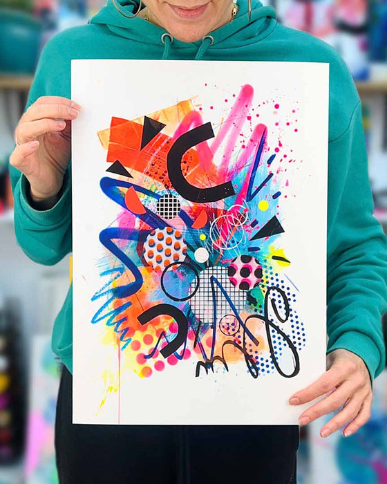 The artist Kate Elkington holding a vibrant, colourful abstract artwork featuring bold, graphic shapes, dynamic brushstrokes, and playful patterns. The composition includes splashes of neon pink, orange, and blue, layered with geometric elements such as dots, grids, and curved black lines, radiating energy and movement.