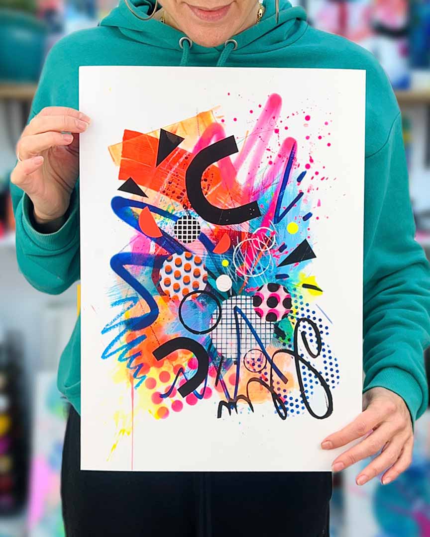 The artist Kate Elkington holding a vibrant, colourful abstract artwork featuring bold, graphic shapes, dynamic brushstrokes, and playful patterns. The composition includes splashes of neon pink, orange, and blue, layered with geometric elements such as dots, grids, and curved black lines, radiating energy and movement.