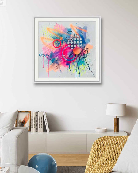 An interior with a white framed vibrant abstract artwork by Kate Elkington bursting with neon hues of pink, orange, yellow, and blue. The composition features bold circular shapes, polka dot patterns, energetic paint splatters, and expressive brushstrokes. Layered textures add contrast and depth, creating a dynamic piece full of playful movement and joyful energy.