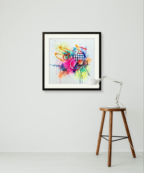 A white interior with an angle-poise lamp on a stool and a black framed vibrant abstract artwork by artist Kate Elkington, bursting with neon hues of pink, orange, yellow, and blue. The composition features bold circular shapes, polka dot patterns, energetic paint splatters, and expressive brushstrokes. Layered textures add contrast and depth, creating a dynamic piece full of playful movement and joyful energy.