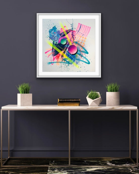 A dark grey interior with a metal framed console table with three small plants on it. Hanging above it is a white framed vibrant abstract artwork  by Kate Elkington, bursting with neon hues of pink, orange, yellow, and blue. The composition features bold circular shapes, polka dot patterns, stripes, energetic paint splatters, and expressive brushstrokes. Layered textures add contrast and depth, creating a dynamic piece full of playful movement and joyful energy.