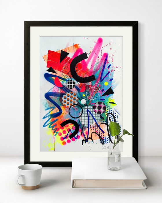 A black framed vibrant, colourful abstract artwork by artist Kate Elkington, leaning on a shelf with a book and coffee cup, featuring bold, graphic shapes, dynamic brushstrokes, and playful patterns. The composition includes splashes of neon pink, orange, bright red, and blue, layered with geometric elements such as dots, grids, and curved black lines, radiating energy and movement.