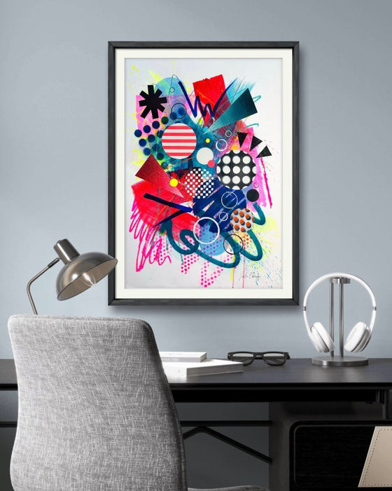 An interior with a desk, lamp and grey study chair and a black framed large, A1 sized, vibrant, colourful abstract artwork by artist Kate Elkington on the wall, featuring bold, graphic shapes, dynamic brushstrokes, and playful patterns. The composition includes splashes of neon pink, bright red, orange, and blue, layered with geometric elements such as dots, grids, and curved black lines, radiating energy and movement.