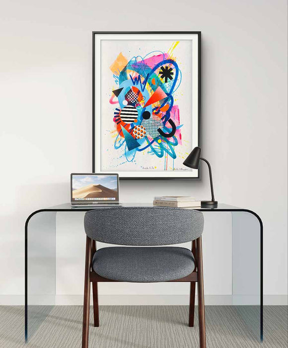 An interior with glass desk and grey chair and a black framed, A1 sized vibrant abstract artwork by Kate Elkington featuring a dynamic mix of bold shapes, patterns, and colors. The composition includes overlapping circles, triangles, and lines in neon pinks, blues, oranges, and yellows, accented with stripes, polka dots, and grid designs. The piece radiates energy and playful creativity.