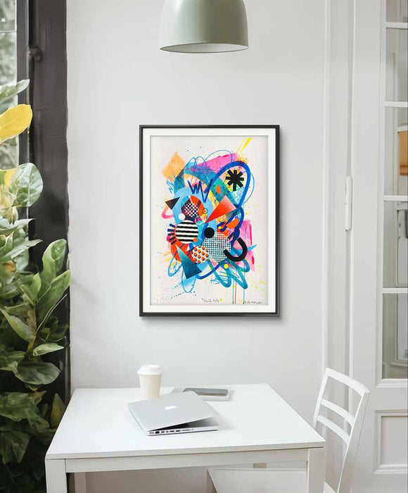 A white interior with a white table, white chair and a black framed A2 sized, vibrant abstract artwork by Kate Elkington featuring a dynamic mix of bold shapes, patterns, and colors. The composition includes overlapping circles, triangles, and lines in neon pinks, blues, oranges, and yellows, accented with stripes, polka dots, and grid designs. The piece radiates energy and playful creativity.