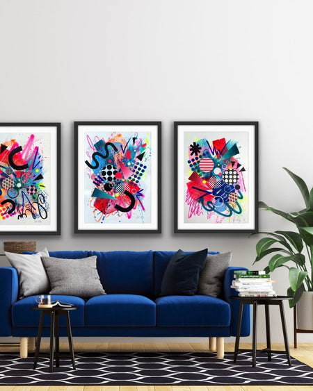 An interior with a blue sofa and plant and a set of three, black framed large, A1 sized, vibrant, colourful abstract artwork's by artist Kate Elkington on the wall, featuring bold, graphic shapes, dynamic brushstrokes, and playful patterns. The composition includes splashes of neon pink, bright red, orange, and blue, layered with geometric elements such as dots, grids, triangles, horseshoe shapes and curved black lines, radiating energy and movement.