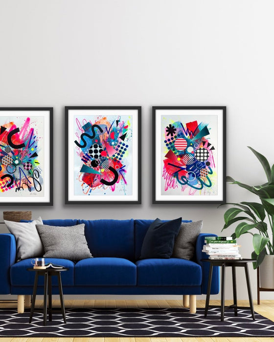 An interior with a blue sofa and plant and a set of three, black framed large, A1 sized, vibrant, colourful abstract artwork's by artist Kate Elkington on the wall, featuring bold, graphic shapes, dynamic brushstrokes, and playful patterns. The composition includes splashes of neon pink, bright red, orange, and blue, layered with geometric elements such as dots, grids, triangles, horseshoe shapes and curved black lines, radiating energy and movement.
