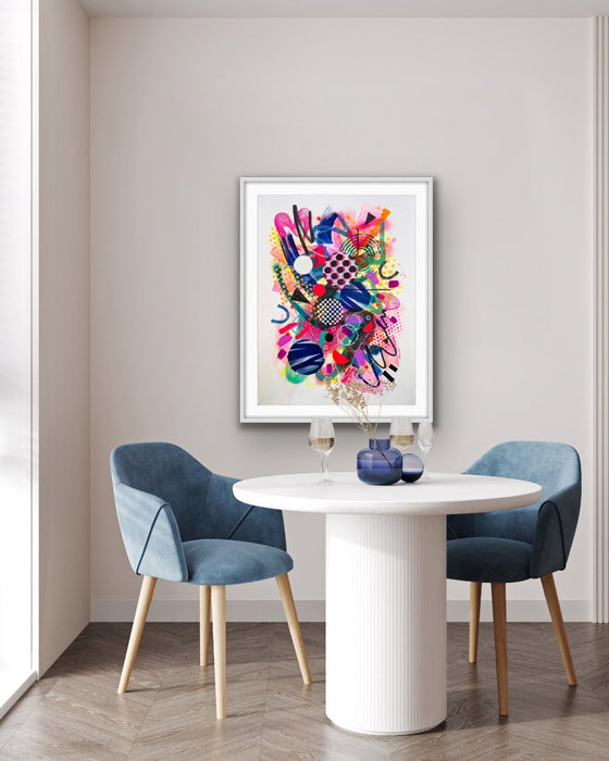 A white interior and blue chairs with a white framed colourful abstract fine art print by Kate Elkington on the wall. Filled with vibrant hues of neon pink, blue, orange, and yellow, the composition of these artworks feature layered circular shapes, bold polka dots, and geometric patterns such as triangles and grids. Energetic brushstrokes and splashes of paint create a lively, dynamic effect, while contrasting tones of black and white add balance. These pieces exude a playful and uplifting vibe.