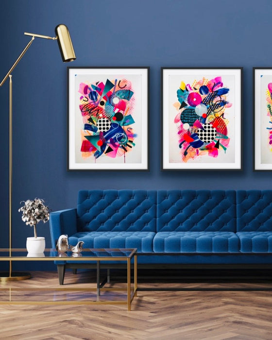 A dark blue interior and blue sofa with three, black framed, colourful abstract fine art prints by Kate Elkington on the wall. Filled with vibrant hues of neon pink, blue, orange, and yellow, the composition of these artworks feature layered circular shapes, bold polka dots, and geometric patterns such as triangles and grids. Energetic brushstrokes and splashes of paint create a lively, dynamic effect, while contrasting tones of black and white add balance. These pieces exude a playful and uplifting vibe.
