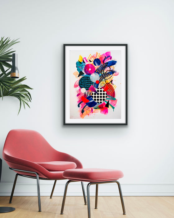 A white interior and pink chair and  with a black framed, colourful abstract fine art print by Kate Elkington on the wall. Filled with vibrant hues of neon pink, blue, orange, and yellow, the composition of the artwork feature layered circular shapes, bold polka dots, and geometric patterns such as triangles and grids. Energetic brushstrokes and splashes of paint create a lively, dynamic effect, while contrasting tones of black and white add balance. This piece exude a playful and uplifting vibe.