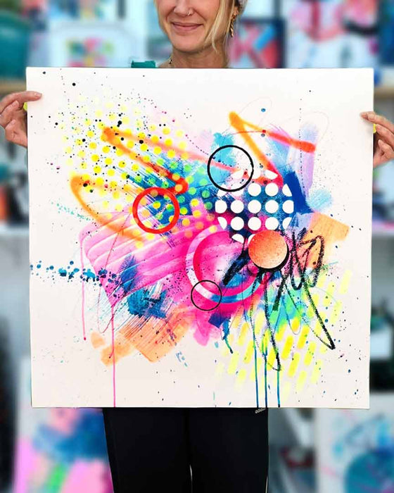 The artist Kate Elkington holding a  vibrant abstract artwork bursting with neon hues of pink, orange, yellow, and blue. The composition features bold circular shapes, polka dot patterns, energetic paint splatters, and expressive brushstrokes. Layered textures add contrast and depth, creating a dynamic piece full of playful movement and joyful energy.
