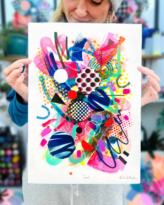The artist Kate Elkington holding an A3 sized colourful abstract fine art print filled with vibrant hues of neon pink, blue, orange, and yellow. The composition features layered circular shapes, bold polka dots, and geometric patterns such as triangles and grids. Energetic brushstrokes and splashes of paint create a lively, dynamic effect, while contrasting tones of black and white add balance. The piece exudes a playful and uplifting vibe.