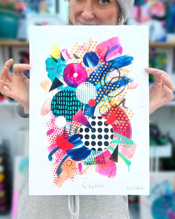 The artist Kate Elkington holding an A3 sized, colourful abstract fine art print filled with vibrant hues of neon pink, blue, orange, and yellow. The composition features layered circular shapes, bold polka dots, and geometric patterns such as triangles and grids. Energetic brushstrokes and splashes of paint create a lively, dynamic effect, while contrasting tones of black and white add balance. The piece exudes a playful and uplifting vibe.