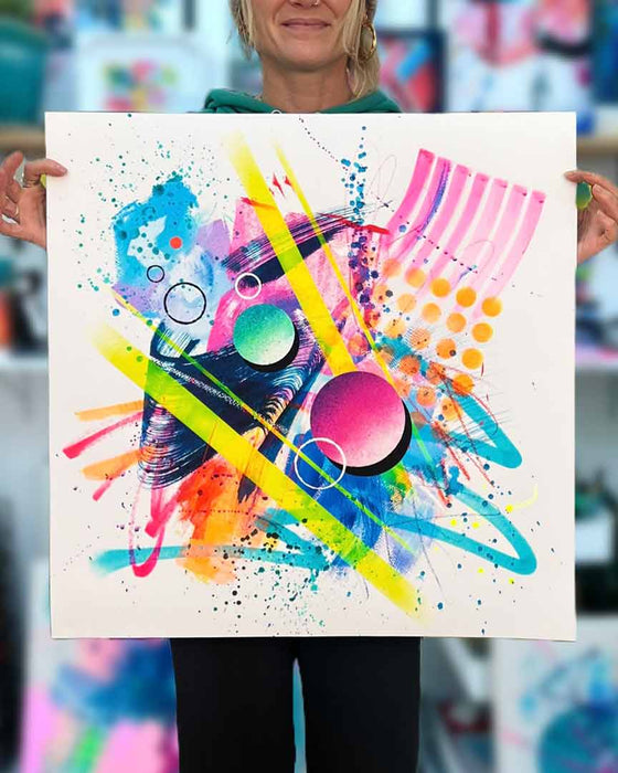 The artist Kate Elkington holding a large 60 by 60cm vibrant abstract artwork bursting with neon hues of pink, orange, yellow, and blue. The composition features bold circular shapes, polka dot patterns, stripes, energetic paint splatters, and expressive brushstrokes. Layered textures add contrast and depth, creating a dynamic piece full of playful movement and joyful energy.