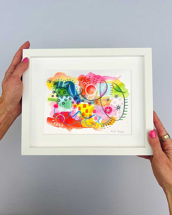 The artist Kate Elkington's hands holding a small, white, framed vibrant abstract giclée art print by Kate Elkington, featuring a playful mix of shapes and colours. Bright yellows, reds, blues, greens, and pinks are blended with soft washes of watercolour, overlaid with whimsical patterns like dots, stars, and swirls. Free-flowing lines and dynamic brushstrokes create a sense of movement, while geometric elements such as squares and circles add structure. The overall composition uplifting and joyful