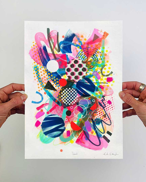 The artist Kate Elkington's hands holding an A4 sized, colourful abstract fine art print filled with vibrant hues of neon pink, blue, orange, and yellow. The composition features layered circular shapes, bold polka dots, and geometric patterns such as triangles and grids. Energetic brushstrokes and splashes of paint create a lively, dynamic effect, while contrasting tones of black and white add balance. The piece exudes a playful and uplifting vibe.