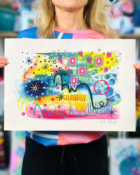 The artist Kate Elkington holding an A3 sized vibrant abstract giclée art print in landscape orientation, featuring a playful mix of shapes and colours. Goldnen yellows, blues, indigo and pinks are blended with soft washes of watercolour, overlaid with whimsical patterns like dots, stars, and swirls. Free-flowing lines and dynamic brushstrokes create a sense of movement, while geometric elements such as squares and circles add structure. The overall composition is uplifting and joyful.
