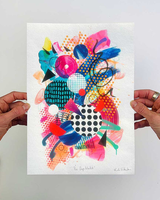 Hands holding an A4 sized, colourful abstract fine art print by Kate Elkington, filled with vibrant hues of neon pink, blue, orange, and yellow. The composition features layered circular shapes, bold polka dots, and geometric patterns such as triangles and grids. Energetic brushstrokes and splashes of paint create a lively, dynamic effect, while contrasting tones of black and white add balance. The piece exudes a playful and uplifting vibe.