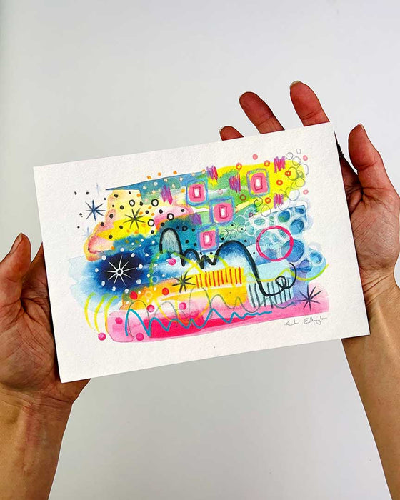 The hands of artist Kate Elkington holding an A5 sized vibrant abstract giclée art print in landscape orientation, featuring a playful mix of shapes and colours. Golden yellows, blues, indigo and pinks blended with soft washes of watercolour, overlaid with whimsical patterns like dots, stars, and swirls. Free-flowing lines and dynamic brushstrokes create a sense of movement, while geometric elements such as squares and circles add structure. The overall composition is uplifting and joyful.