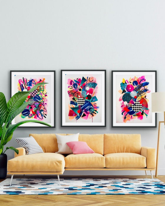 A white interior and yellow sofa with three, black framed, colourful abstract fine art prints by Kate Elkington on the wall. Filled with vibrant hues of neon pink, blue, orange, and yellow, the composition of these artworks feature layered circular shapes, bold polka dots, and geometric patterns such as triangles and grids. Energetic brushstrokes and splashes of paint create a lively, dynamic effect, while contrasting tones of black and white add balance. These pieces exude a playful and uplifting vibe.