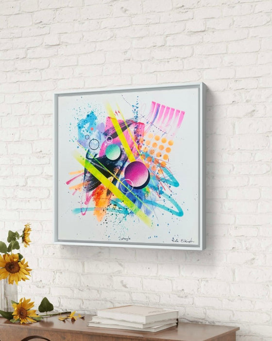 A white, brick wall with a white framed vibrant abstract artwork by Kate Elkington, bursting with neon hues of pink, orange, yellow, and blue. The composition features bold circular shapes, polka dot patterns, stripes, energetic paint splatters, and expressive brushstrokes. Layered textures add contrast and depth, creating a dynamic piece full of playful movement and joyful energy.