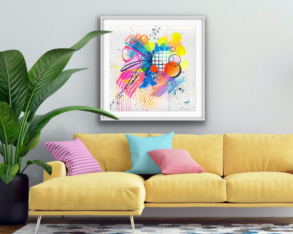 A framed colourful abstract artwork by Kate Elkington featuring expressive textures, mark making and bold graphic shapes in pinks, blues, turquoise, neon orange, neon yellow and neon pink in a room with a yellow sofa, colourful cusions and a plant