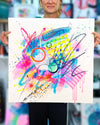 The artist Kate Elkington holding a large 60 by 60cm dynamic abstract artwork featuring bold splashes of neon pink, jade green, yellow, orange, and blue, combined with graphic circle shapes, stripes, black accents and white grid patterns. Expressive brush strokes, crayon scribbles and paint drips add movement and energy, creating a vibrant and playful composition that radiates joy and positivity.