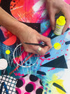 A close up of the hands of artist Kate Elkington hand-finishing a limited edition fine art giclée print with a pop of neon fluorescent yellow paint on some small circular shapes. The rest of the print depicts layers of texture, vibrant blues and pinks and bold graphic shapes such as triangles. semi circles and horseshoe shapes.