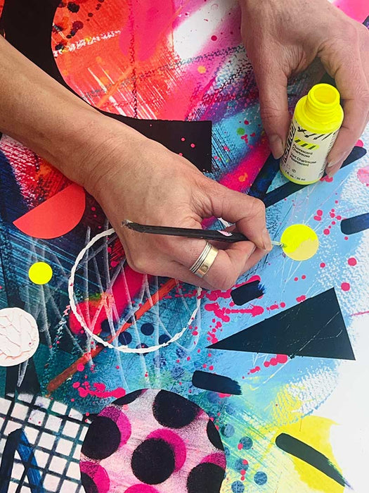 A close up of the hands of artist Kate Elkington hand-finishing a limited edition fine art giclée print with a pop of neon fluorescent yellow paint on some small circular shapes. The rest of the print depicts layers of texture, vibrant blues and pinks and bold graphic shapes such as triangles. semi circles and horseshoe shapes.