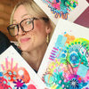 Artist Kate Elkington holding her colourful abstract 'Happy Doodle' art pieces with a blurred background