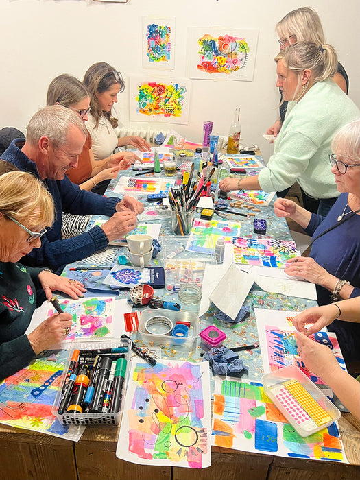 Group of people engaged in a craft or art activity at a table with various materials and colorful artwork.
