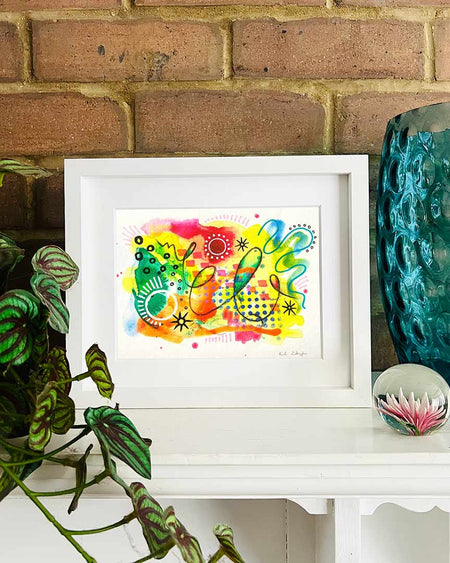 A small. white, framed, vibrant abstract giclée art print by Kate Elkington sitting on a white mantle with blue vase and plant. the artwork features a playful mix of shapes and colours. Bright yellows, oranges, blues, greens, and hot pinks are blended with soft washes of watercolour, overlaid with whimsical patterns like dots, stars, and swirls. Free-flowing lines and dynamic brushstrokes create a sense of movement, while geometric elements such as squares and circles add structure. 