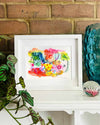 A small, white, framed vibrant abstract giclée art print by Kate Elkington sitting on a white mantle with blue vase and plant. The artwork features a playful mix of shapes and colours. Bright yellows, reds, blues, greens, and pinks are blended with soft washes of watercolour, overlaid with whimsical patterns like dots, stars, and swirls. Free-flowing lines and dynamic brushstrokes create a sense of movement, while geometric elements such as squares and circles add structure. 
