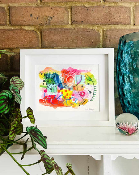 A small, white, framed vibrant abstract giclée art print by Kate Elkington sitting on a white mantle with blue vase and plant. The artwork features a playful mix of shapes and colours. Bright yellows, reds, blues, greens, and pinks are blended with soft washes of watercolour, overlaid with whimsical patterns like dots, stars, and swirls. Free-flowing lines and dynamic brushstrokes create a sense of movement, while geometric elements such as squares and circles add structure. 