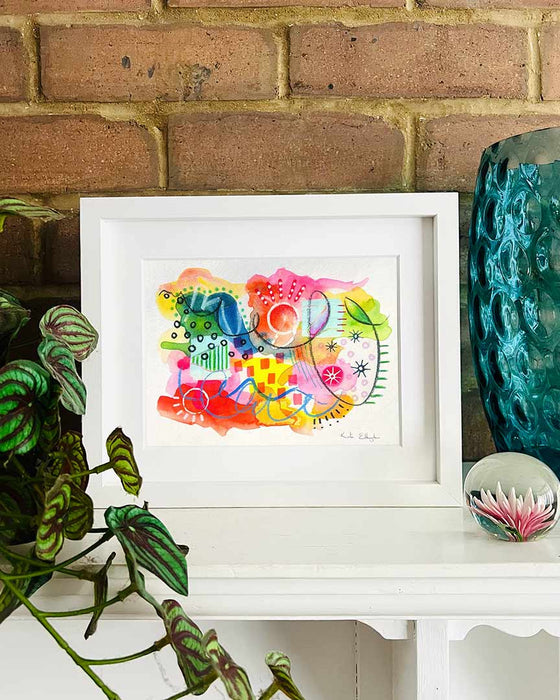 A small, white, framed vibrant abstract giclée art print by Kate Elkington sitting on a white mantle with blue vase and plant. The artwork features a playful mix of shapes and colours. Bright yellows, reds, blues, greens, and pinks are blended with soft washes of watercolour, overlaid with whimsical patterns like dots, stars, and swirls. Free-flowing lines and dynamic brushstrokes create a sense of movement, while geometric elements such as squares and circles add structure. 