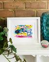 A small, white, framed vibrant abstract giclée art print by Kate Elkington sitting on a white mantle along with a blue glass vase and a plant. The artwork features a playful mix of shapes and colours. Golden yellows, blues, indigo and pinks are blended with soft washes of watercolour, overlaid with whimsical patterns like dots, stars, and swirls. Free-flowing lines and dynamic brushstrokes create a sense of movement, while geometric elements such as squares and circles add structure. 