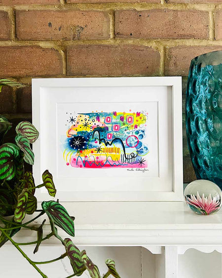 A small, white, framed vibrant abstract giclée art print by Kate Elkington sitting on a white mantle along with a blue glass vase and a plant. The artwork features a playful mix of shapes and colours. Golden yellows, blues, indigo and pinks are blended with soft washes of watercolour, overlaid with whimsical patterns like dots, stars, and swirls. Free-flowing lines and dynamic brushstrokes create a sense of movement, while geometric elements such as squares and circles add structure. 