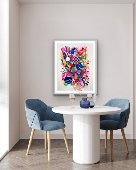 A white interior and blue chairs with a white framed colourful abstract fine art print by Kate Elkington on the wall. Filled with vibrant hues of neon pink, blue, orange, and yellow, the composition of these artworks feature layered circular shapes, bold polka dots, and geometric patterns such as triangles and grids. Energetic brushstrokes and splashes of paint create a lively, dynamic effect, while contrasting tones of black and white add balance. These pieces exude a playful and uplifting vibe.