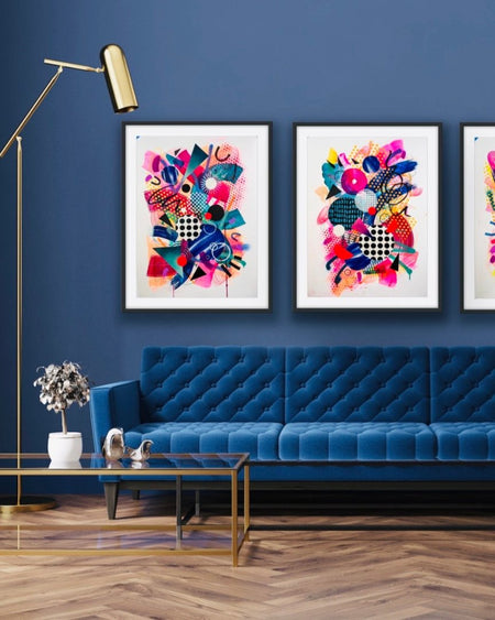 A dark blue interior and blue sofa with three, black framed, colourful abstract fine art prints by Kate Elkington on the wall. Filled with vibrant hues of neon pink, blue, orange, and yellow, the composition of these artworks feature layered circular shapes, bold polka dots, and geometric patterns such as triangles and grids. Energetic brushstrokes and splashes of paint create a lively, dynamic effect, while contrasting tones of black and white add balance. These pieces exude a playful and uplifting vibe.