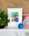 A small, white, framed vibrant abstract giclée art print by Kate Elkington on a white mantle with plant and vase. Featuring a playful mix of shapes and colours. Greens, yellows, blues, indigo and pinks are blended with soft washes of watercolour, overlaid with whimsical patterns like dots, stars, and swirls. Free-flowing lines and dynamic brushstrokes create a sense of movement, while geometric elements such as squares and circles add structure.