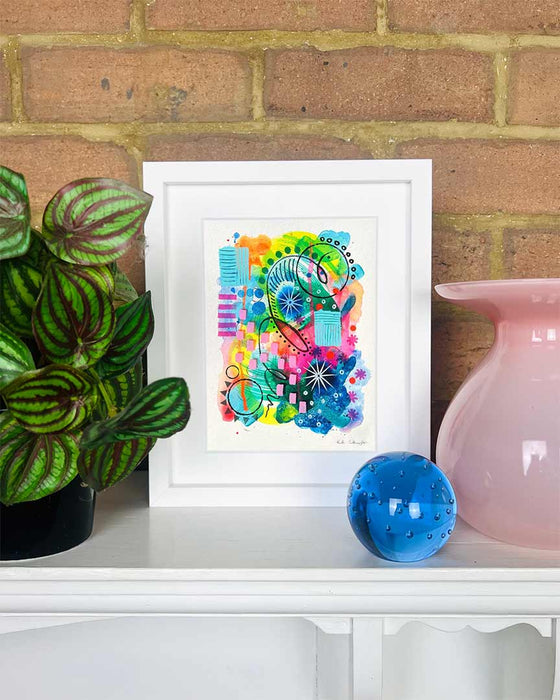 A small, white, framed vibrant abstract giclée art print by Kate Elkington on a white mantle with plant and vase. Featuring a playful mix of shapes and colours. Greens, yellows, blues, indigo and pinks are blended with soft washes of watercolour, overlaid with whimsical patterns like dots, stars, and swirls. Free-flowing lines and dynamic brushstrokes create a sense of movement, while geometric elements such as squares and circles add structure.