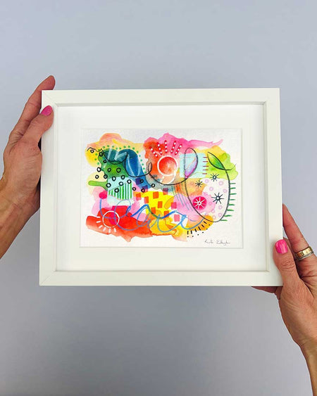The artist Kate Elkington's hands holding a small, white, framed vibrant abstract giclée art print by Kate Elkington, featuring a playful mix of shapes and colours. Bright yellows, reds, blues, greens, and pinks are blended with soft washes of watercolour, overlaid with whimsical patterns like dots, stars, and swirls. Free-flowing lines and dynamic brushstrokes create a sense of movement, while geometric elements such as squares and circles add structure. The overall composition uplifting and joyful