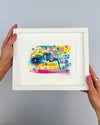 The artist Kate Elkington's hands holding a small, white, framed vibrant abstract giclée art print, featuring a playful mix of shapes and colours. Golden yellows, blues, indigo and pinks are blended with soft washes of watercolour, overlaid with whimsical patterns like dots, stars, and swirls. Free-flowing lines and dynamic brushstrokes create a sense of movement, while geometric elements such as squares and circles add structure. The overall composition uplifting and joyful
