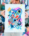 The artist Kate Elkington holding a vibrant abstract artwork featuring a dynamic mix of bold shapes, patterns, and colours. The composition includes overlapping circles, triangles, and lines in neon pinks, blues, oranges, and yellows, accented with stripes, polka dots, and grid designs. The piece radiates energy and playful creativity.
