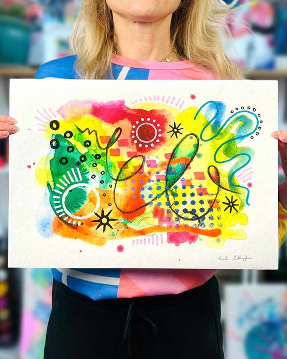 The artist Kate Elkington holding an A3 sized vibrant abstract giclée art print in landscape orientation, featuring a playful mix of shapes and colours. Bright yellows, oranges, blues, greens, and hot pinks are blended with soft washes of watercolour, overlaid with whimsical patterns like dots, stars, and swirls. Free-flowing lines and dynamic brushstrokes create a sense of movement, while geometric elements such as squares and circles add structure. The overall composition is uplifting and joyful.