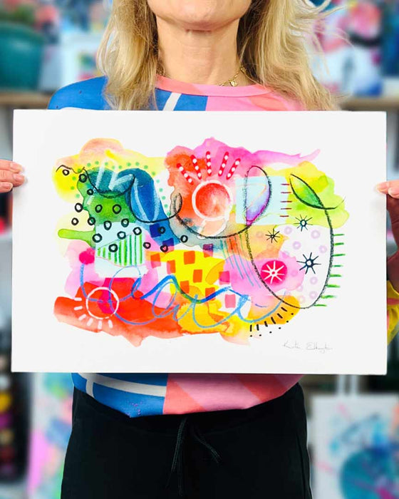 The artist Kate Elkington holding an A3 sized vibrant abstract giclée art print in landscape orientation, featuring a playful mix of shapes and colours. Bright yellows, reds, blues, greens, and pinks are blended with soft washes of watercolour, overlaid with whimsical patterns like dots, stars, and swirls. Free-flowing lines and dynamic brushstrokes create a sense of movement, while geometric elements such as squares and circles add structure. The overall composition is uplifting and joyful.