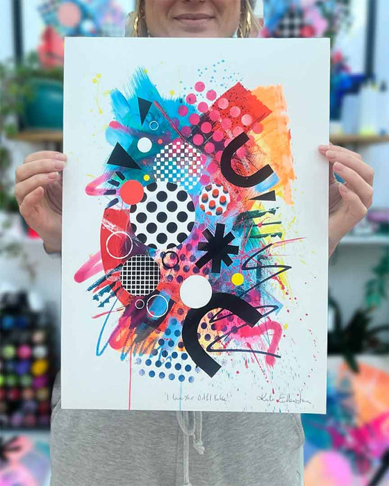 The artist Kate Elkington holding an A3 sized, vibrant, colourful abstract artwork featuring bold, graphic shapes, dynamic brushstrokes, and playful patterns. The composition includes splashes of neon pink, orange, and blue, layered with geometric elements such as dots, grids, and curved black lines, radiating energy and movement.